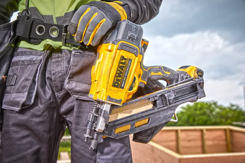 Personne tenant un cloueur DEWALT DCN930N, portant des gants et un équipement sur un chantier extérieur.