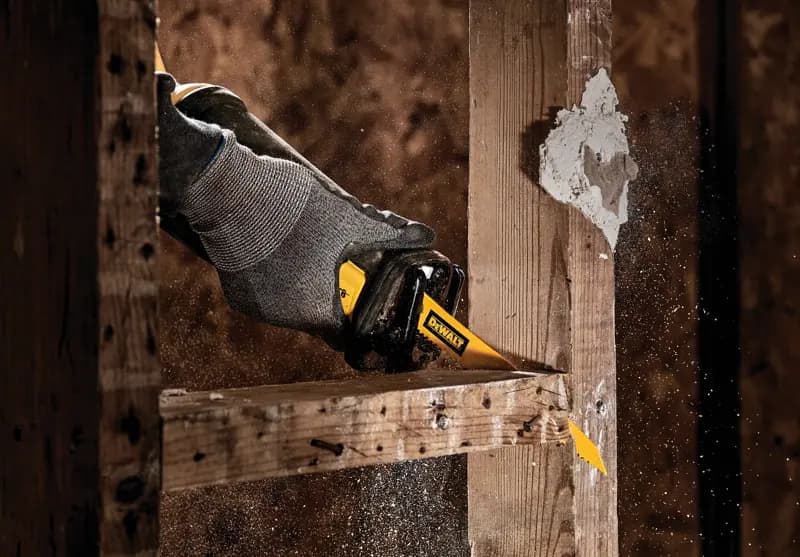 Gros plan sur une main portant un gant gris utilisant une scie sabre DEWALT pour couper une poutre en bois, avec de la sciure projetée dans l'air.