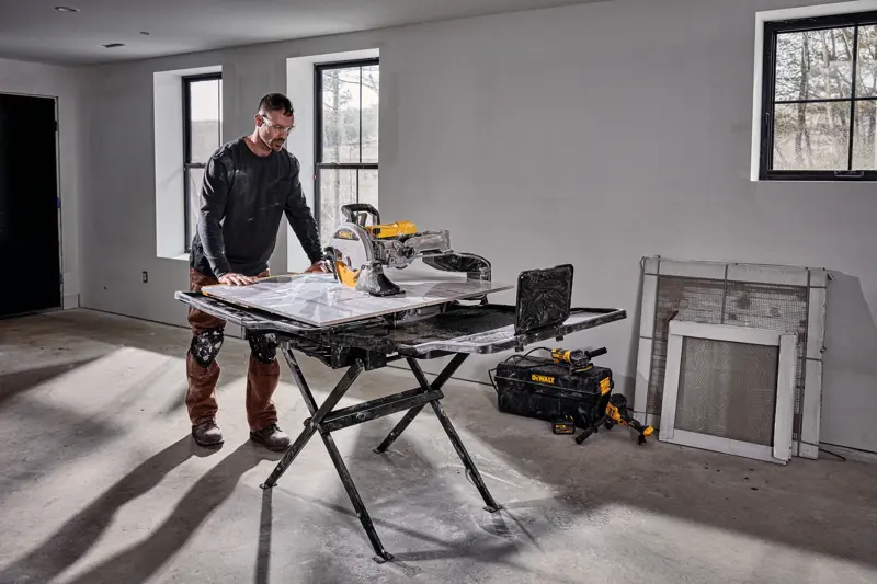 Une personne se tient à côté d’une scie à carrelage DEWALT D36000S installée sur son support dans une pièce en cours de construction. La scie est placée sur un support pliable noir, avec des outils et un sac à outils DEWALT au sol à proximité. La pièce possède des murs non finis, des fenêtres et des matériaux de construction appuyés contre le mur.