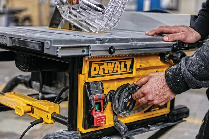 Gros plan d'une personne ajustant les réglages sur une scie à table DEWALT, avec un panneau de commande jaune comportant des interrupteurs et des cadrans, dans un atelier.