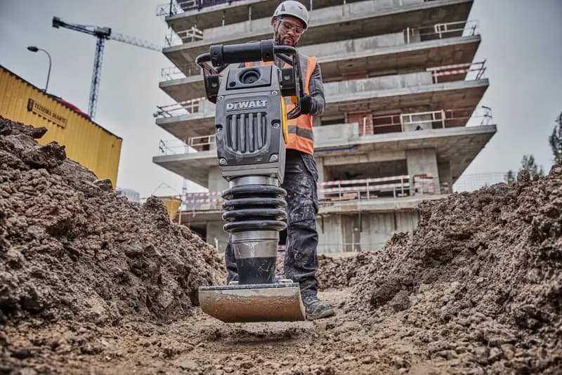 Compacteur DEWALT utilisé sur un chantier, opéré par un ouvrier équipé, entouré de terre et de bâtiments.