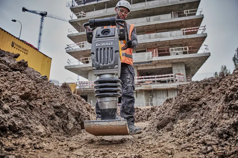 Compacteur DEWALT utilisé sur un chantier, opéré par un ouvrier équipé, entouré de terre et de bâtiments.